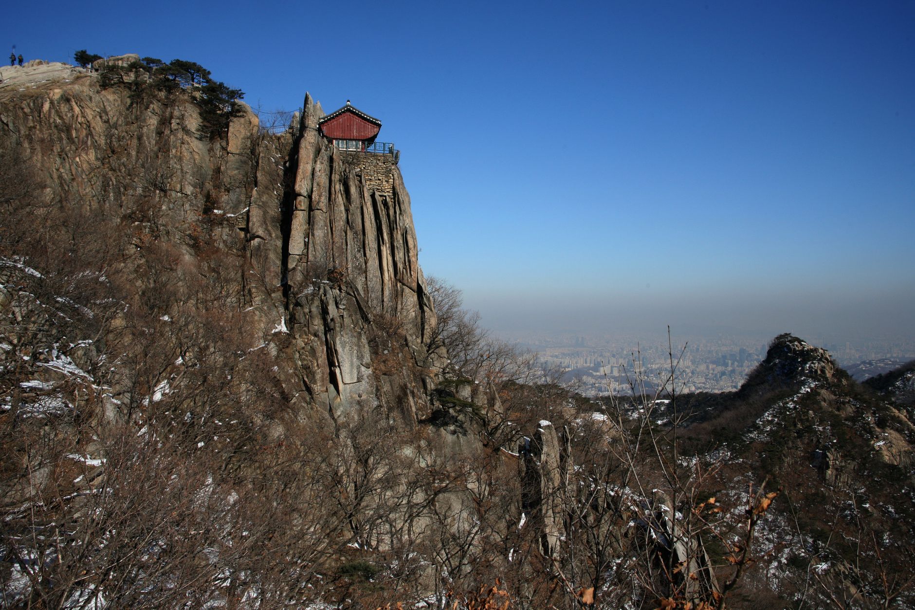 A rocky cliff with a traditional structure and scenic city view under a clear blue sky, captured in winter.