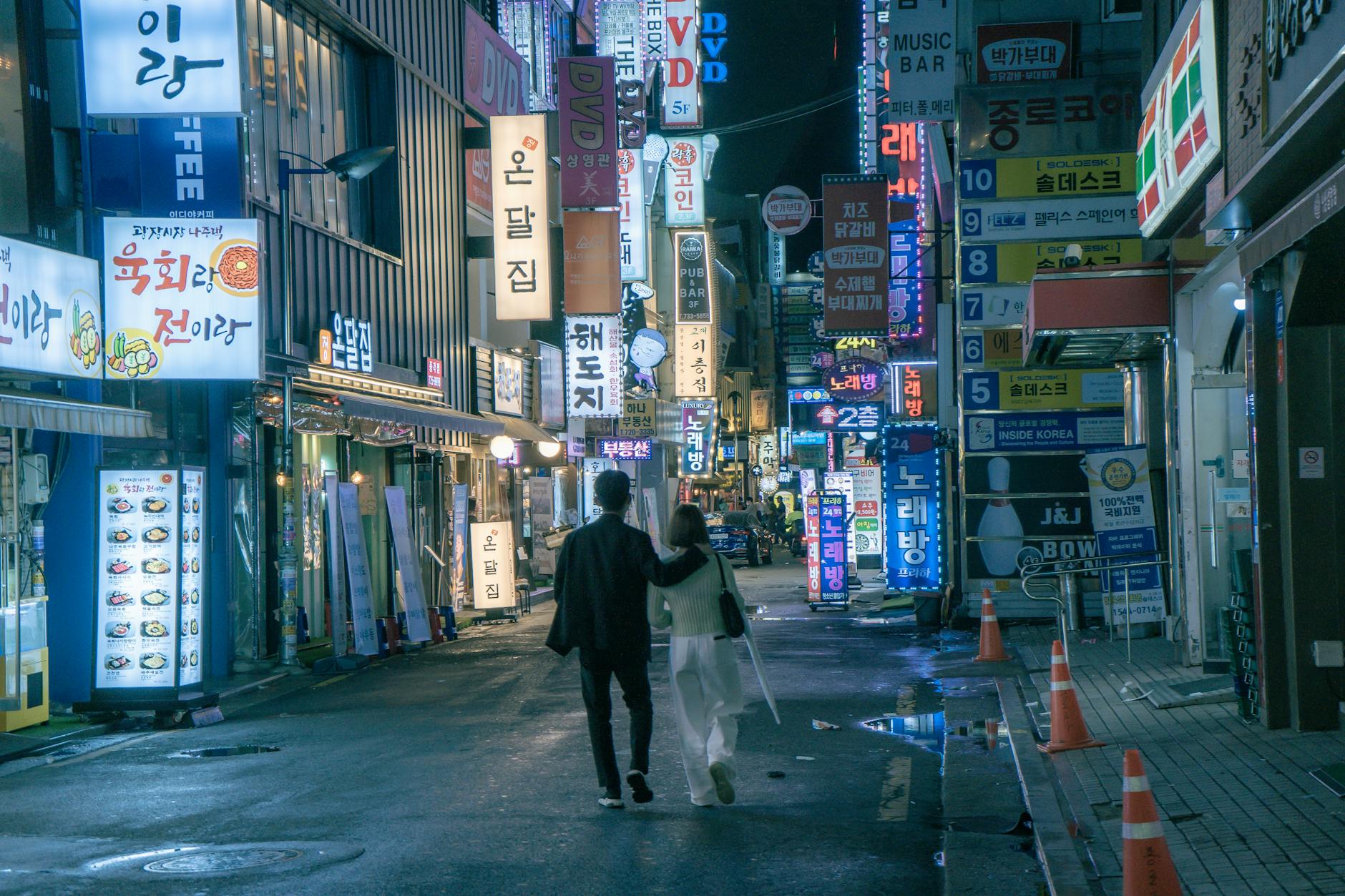 An Asian couple walks through the vibrant, neon-lit streets of Seoul at night, surrounded by shops and signs.
