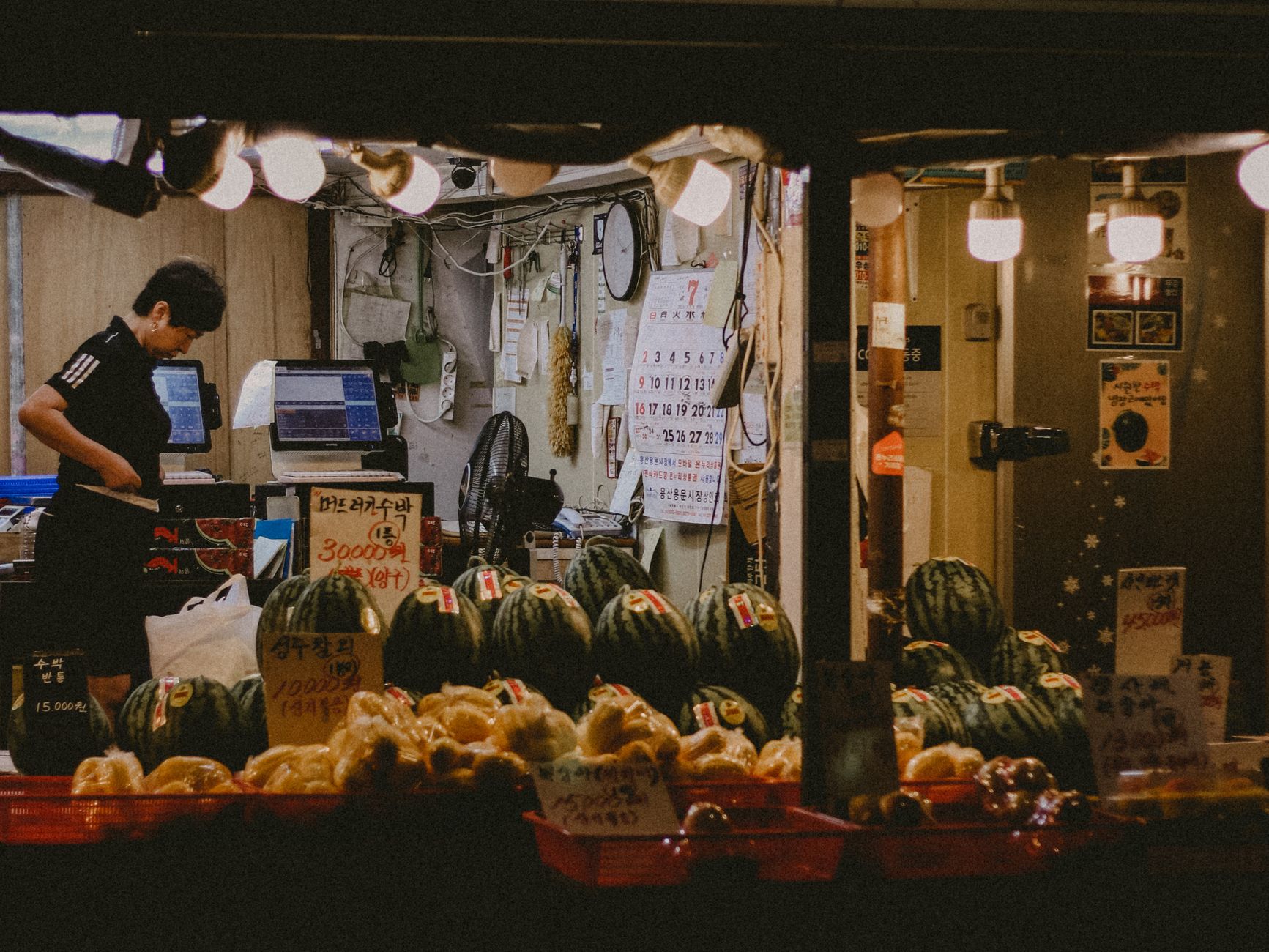 A vendor in an illuminated night market sells watermelons and fresh produce.