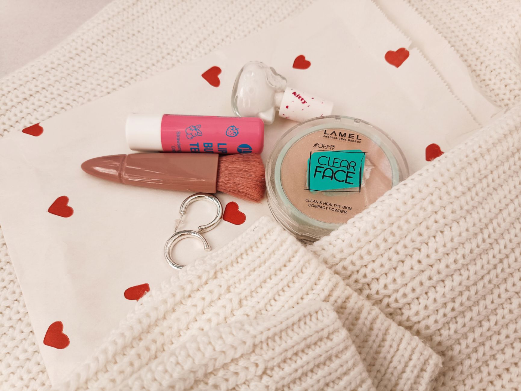 Stylish flat lay featuring makeup items, white knit, and heart-patterned cloth for beauty inspiration.