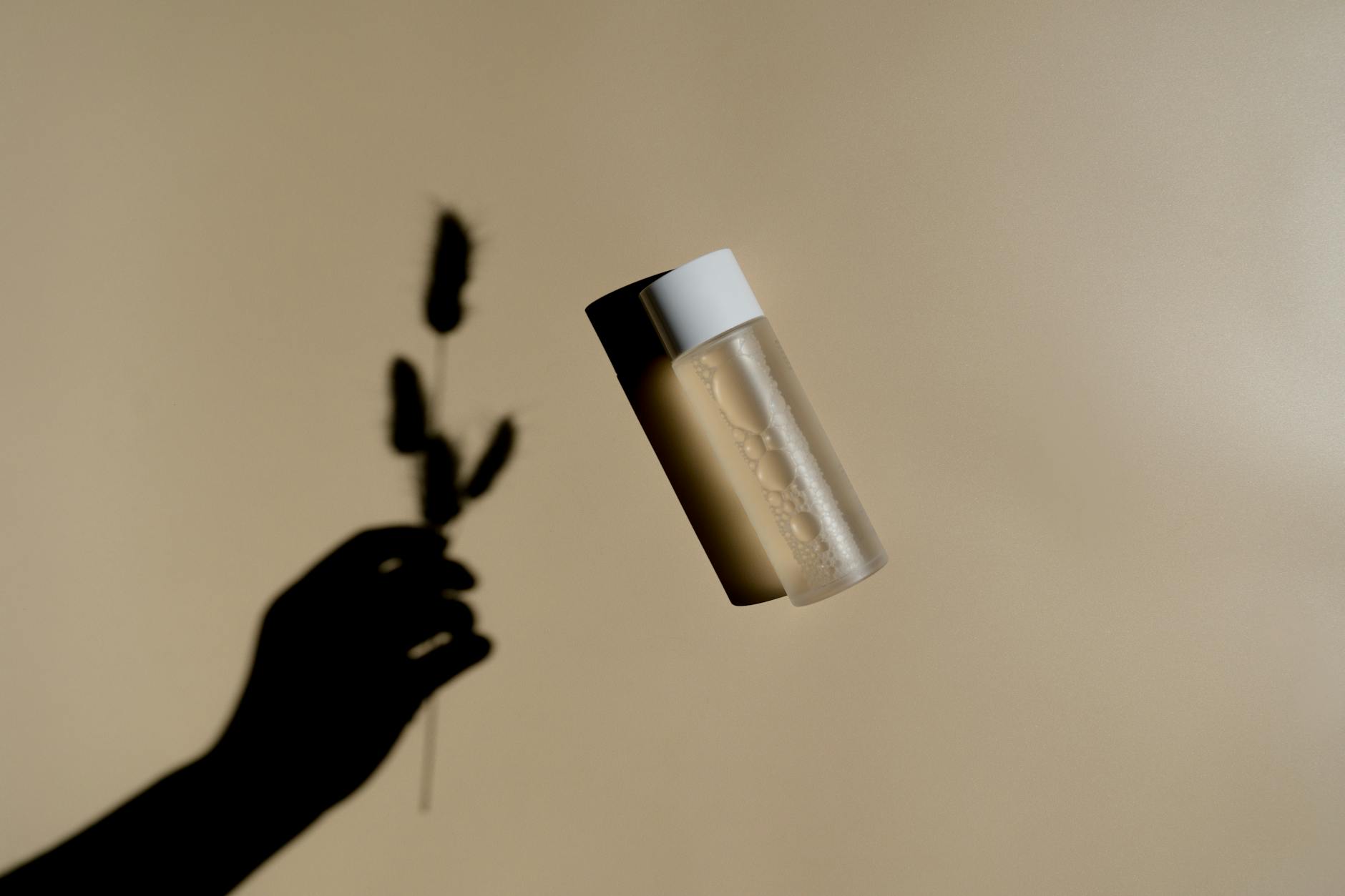 An artistic composition showcasing a shadow of a hand holding a plant beside a suspended bottle on a beige background.