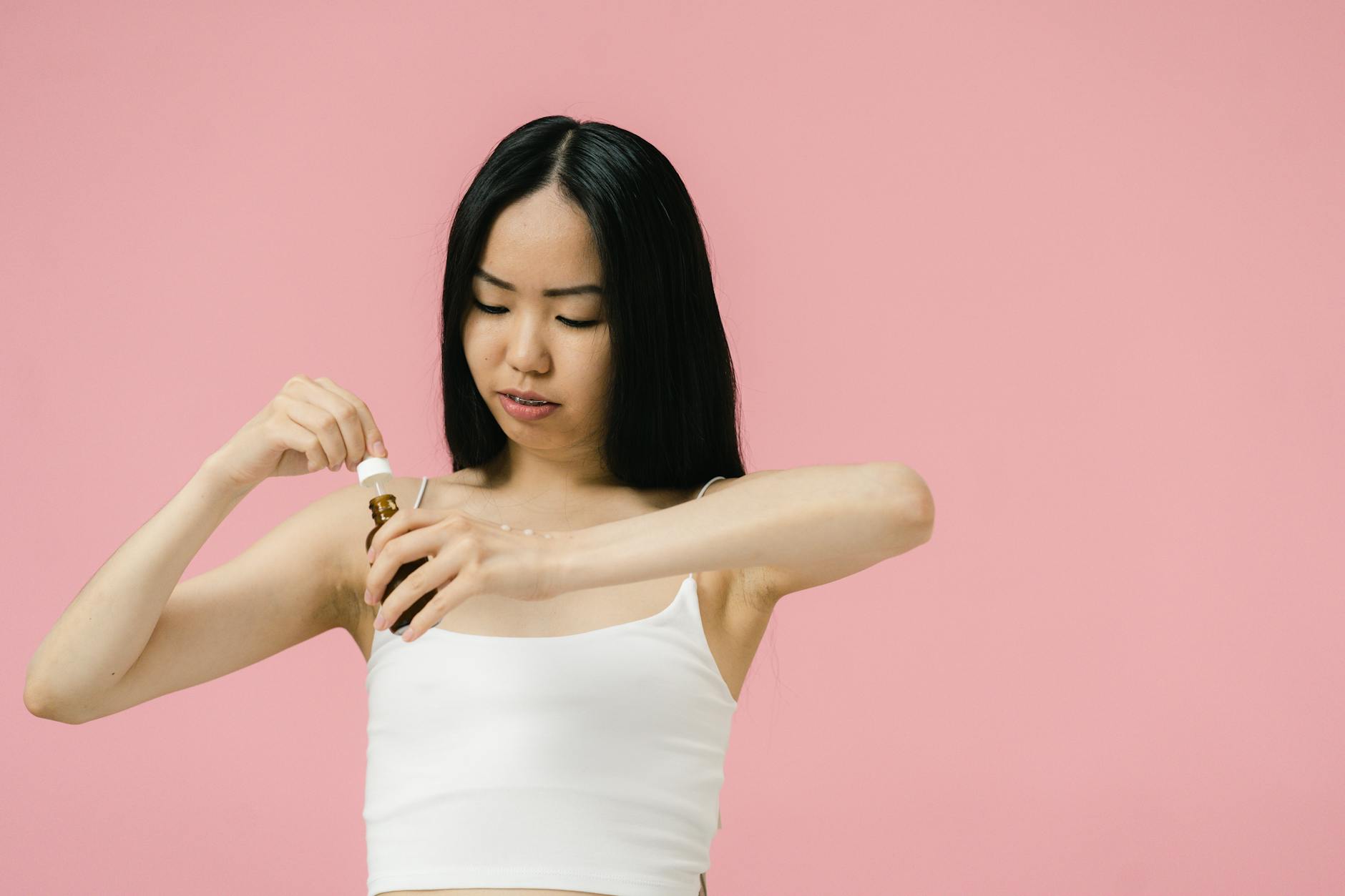 Asian woman using skincare product with pipette against a pink background, promoting self-care and wellness.
