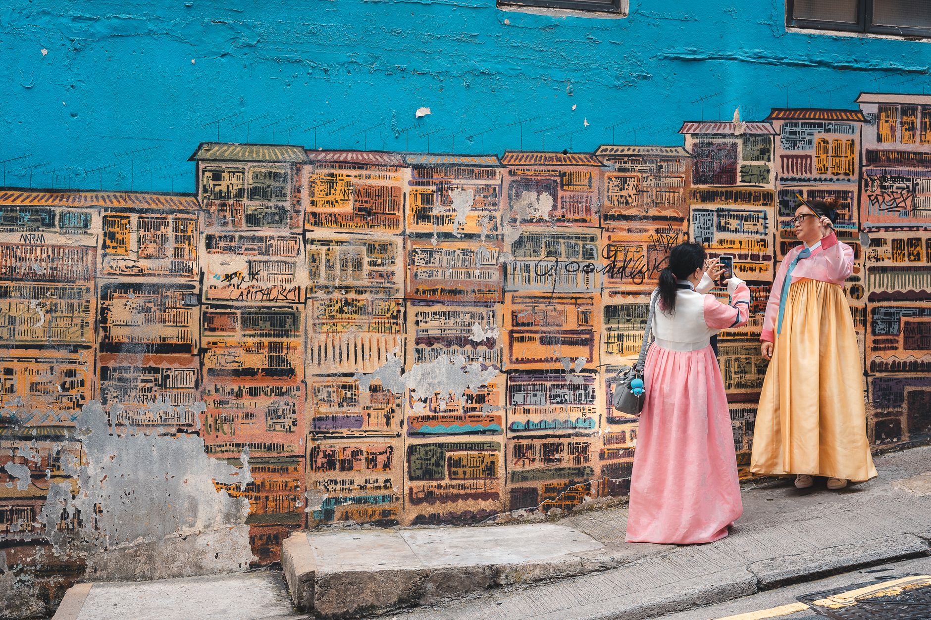 Two women in traditional Korean dresses pose by a vibrant mural in Hong Kong.