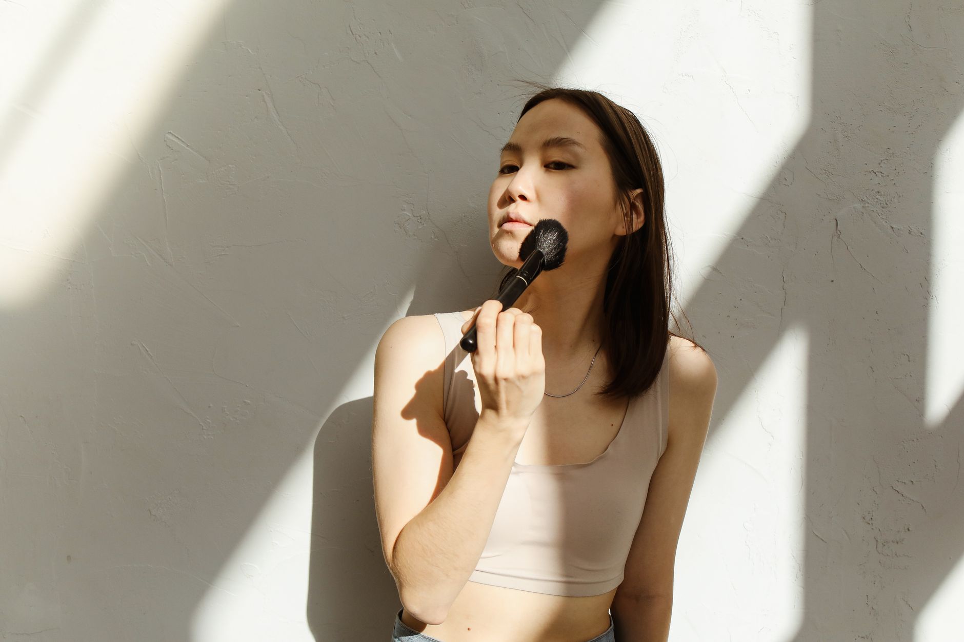 Asian woman using makeup brush on face against a sunlit wall indoors.