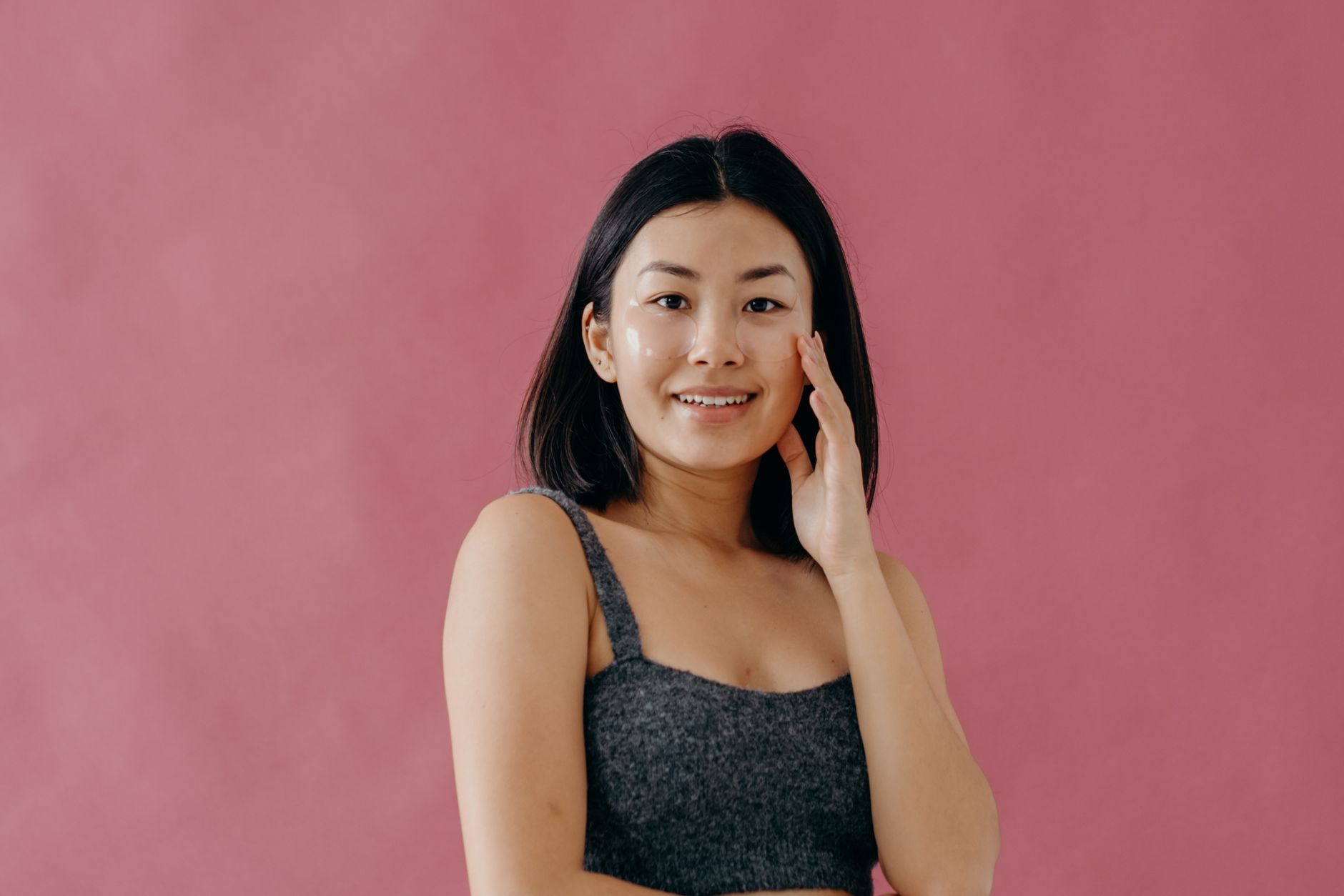 Studio portrait of a smiling Asian woman with under-eye skincare patches against a pink background.